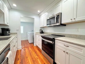 A kitchen with white cabinets, a stainless steel microwave, a stove, and a washing machine in the adjacent room.