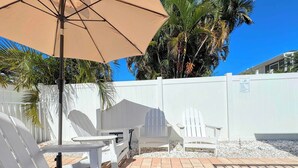 A patio area with Adirondack chairs, a table, and a beige umbrella. - 231 Pearl Island Cottage Retreat w Private Pool (Fort Myers Beach)