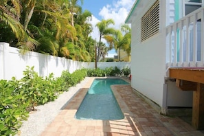 A swimming pool with a white fence, surrounded by greenery and a white building. - 231 Pearl Island Cottage Retreat w Private Pool (Fort Myers Beach)