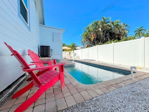 A swimming pool with red Adirondack chairs, a white fence, and a clear blue sky.