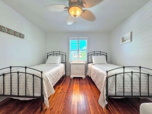Un dormitorio con dos camas individuales, un ventilador de techo, una ventana con persianas y una mesita de noche.