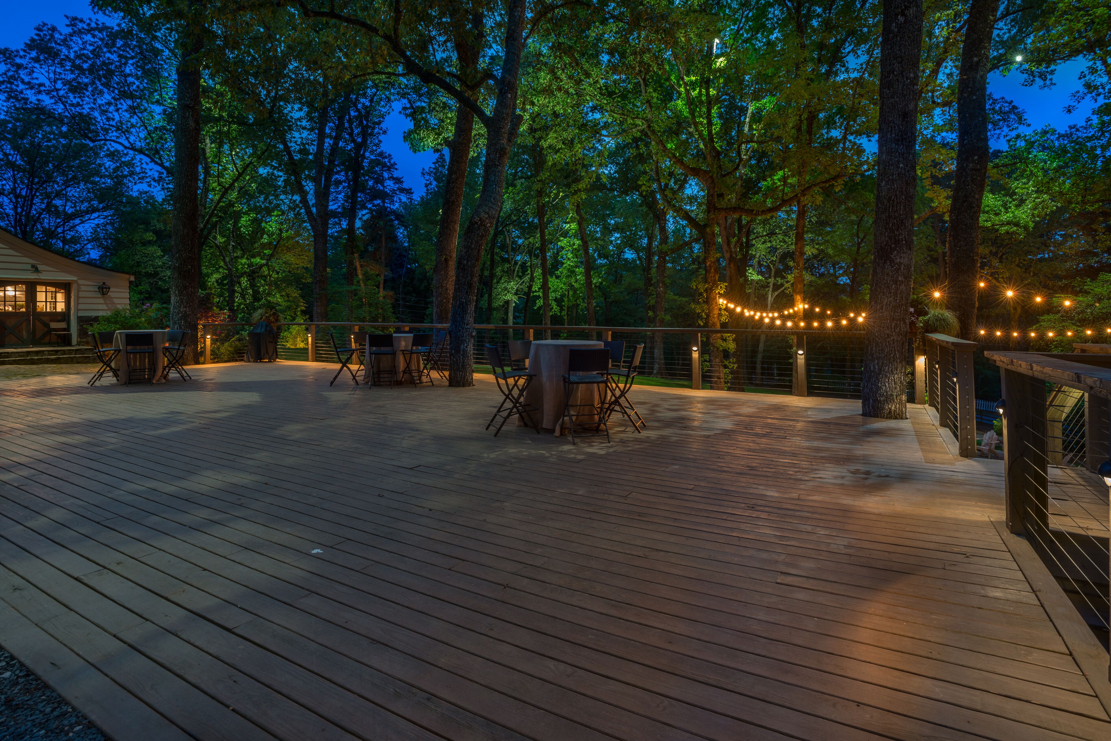 An outdoor deck with wooden flooring, surrounded by trees and string lights.