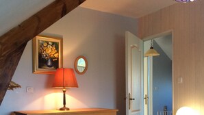 Une chambre à coucher avec une commode en bois, une lampe rouge, un tableau floral encadré et une porte donnant sur une autre pièce.
