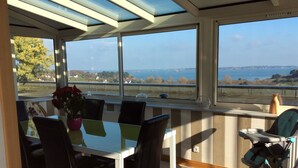 Un espace repas comprenant une table et des chaises, de grandes fenêtres offrant une vue sur la mer et le paysage, et une chaise accessible aux personnes en fauteuil roulant.