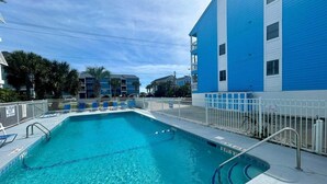 Une piscine entourée d’une clôture blanche, avec des chaises longues bleues, et un bâtiment dont les murs extérieurs sont peints en bleu et en blanc.