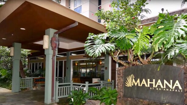Entrada de un hotel con un paseo techado, un letrero que dice "MAHANA AT KA'ANAPALI", y exuberante vegetación tropical.