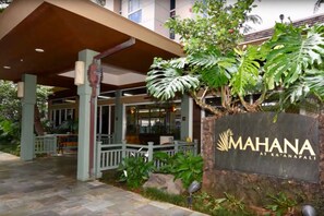 Entrada de un hotel con un paseo techado, un letrero que dice "MAHANA AT KA'ANAPALI", y exuberante vegetación tropical.