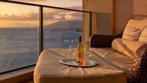 Un balcón con vistas al mar y a las montañas, un sofá con cojines, una bandeja con una botella y copas, y una cesta de mimbre.