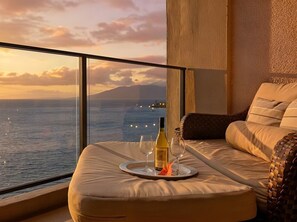 Un balcón con vistas al mar y a las montañas, un sofá con cojines, una bandeja con una botella y copas, y una cesta de mimbre.
