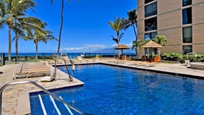 A swimming pool with lounge chairs and umbrellas, surrounded by palm trees and a building with large windows. - ❤️ Renovated 8th Floor Studio at The Mahana!! ❤️ (Lahaina)