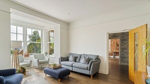 Un salón amplio con un sofá gris, un sillón azul y un reposapiés. Tiene piso de madera, un ventanal grande y una puerta que da a otra habitación.