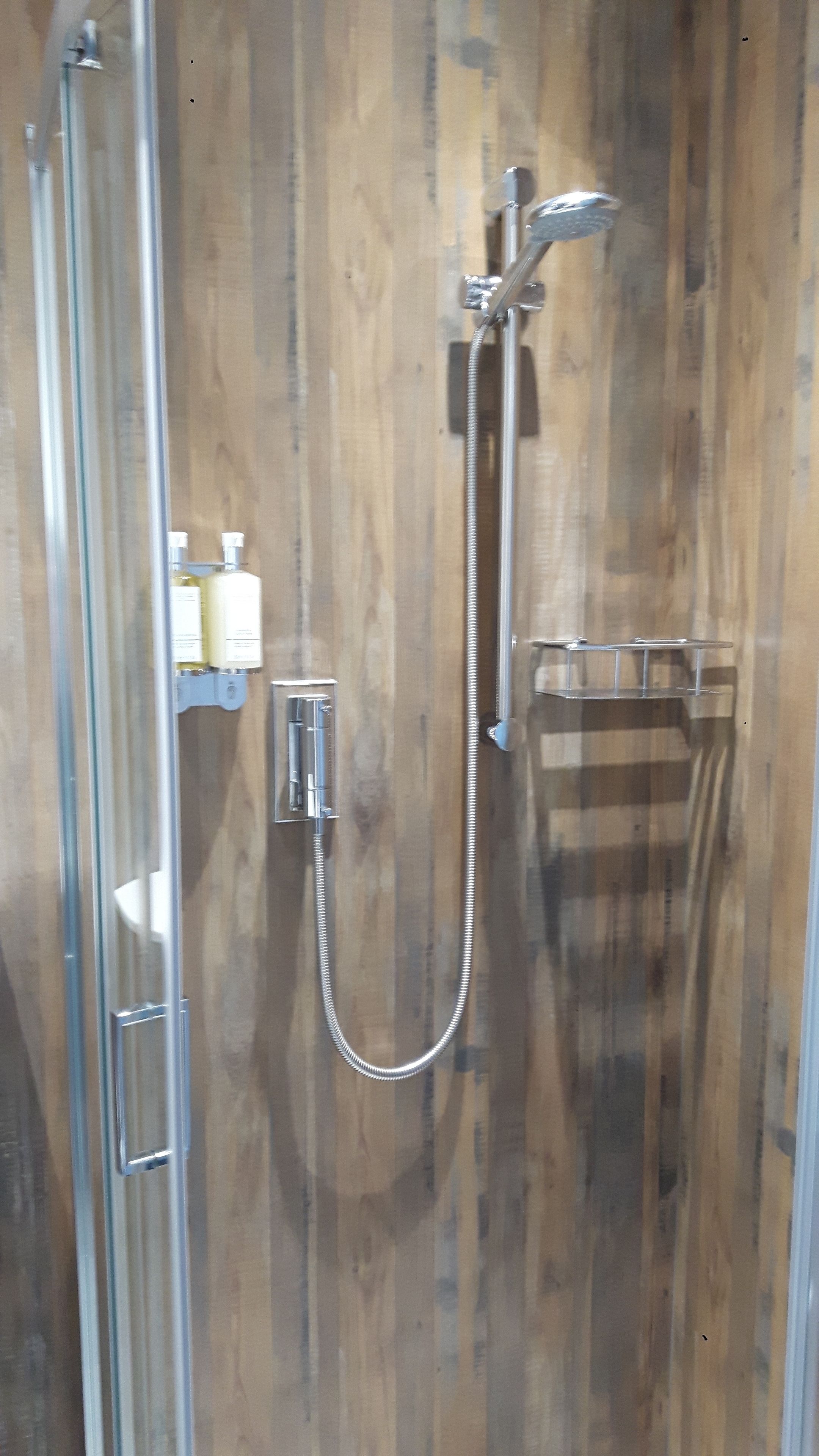 A glass-enclosed shower stall with a wooden wall, a handheld showerhead, a wall-mounted faucet, and a soap dispenser.