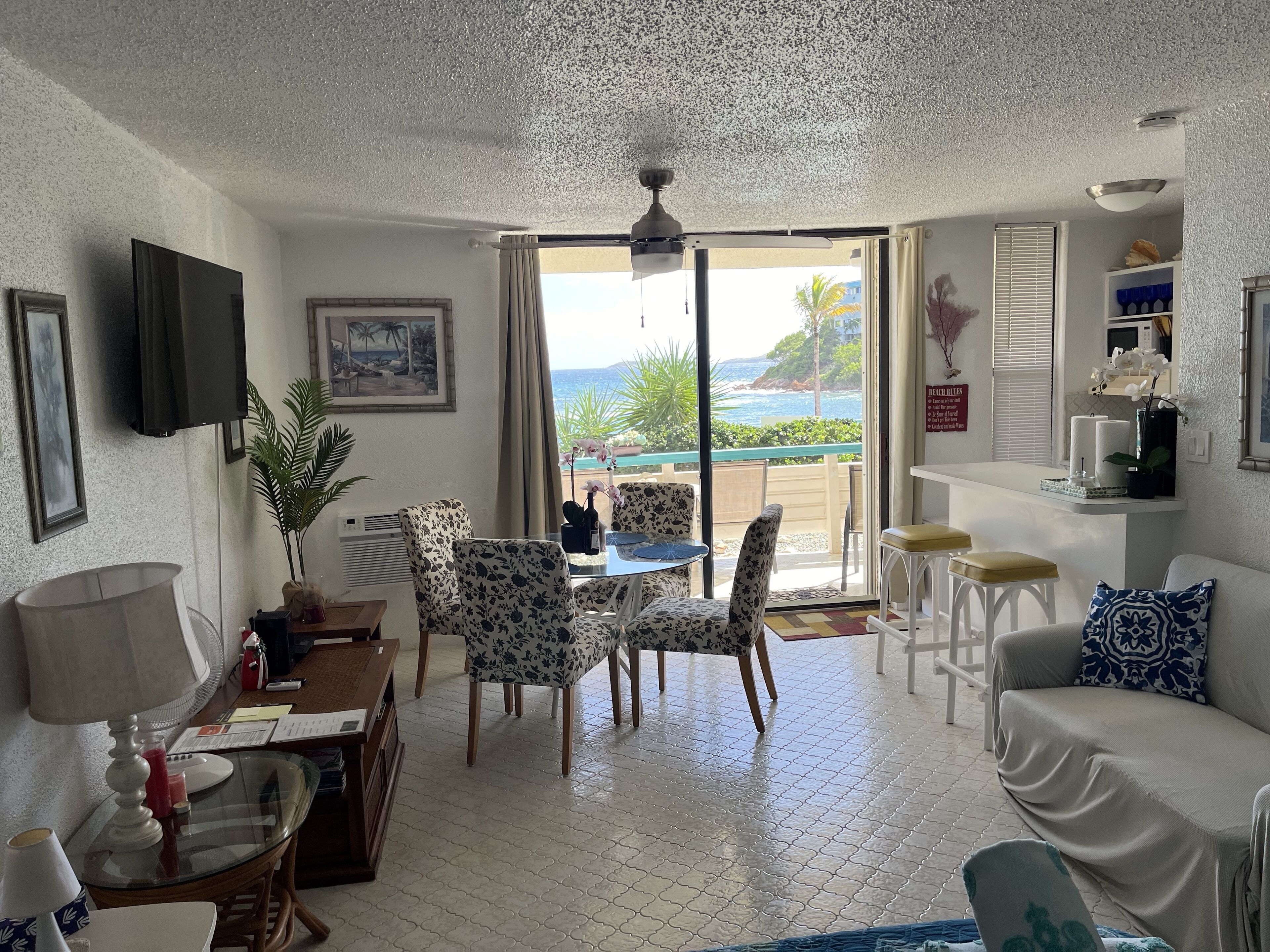 A living room with a dining area, a TV mounted on the wall, a ceiling fan, and a view of the ocean through the open door.