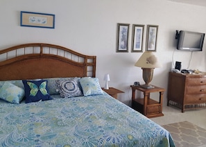 Une chambre à coucher avec une tête de lit en bois, un lit recouvert d’une literie à motifs, une table de chevet avec une lampe, une commode et un téléviseur fixé au mur.