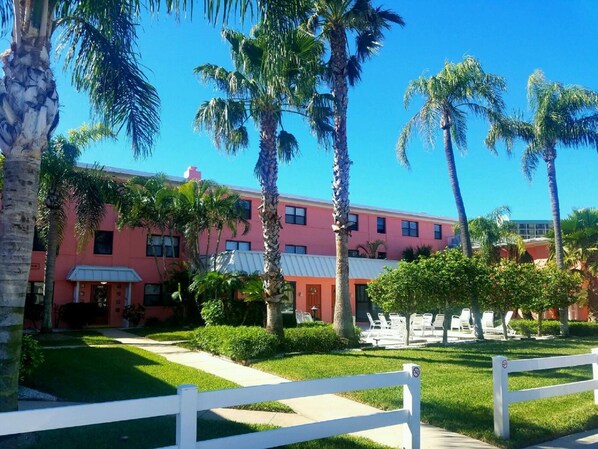 Un edificio rosa con una cerca blanca al frente, rodeado de palmeras y follaje.
