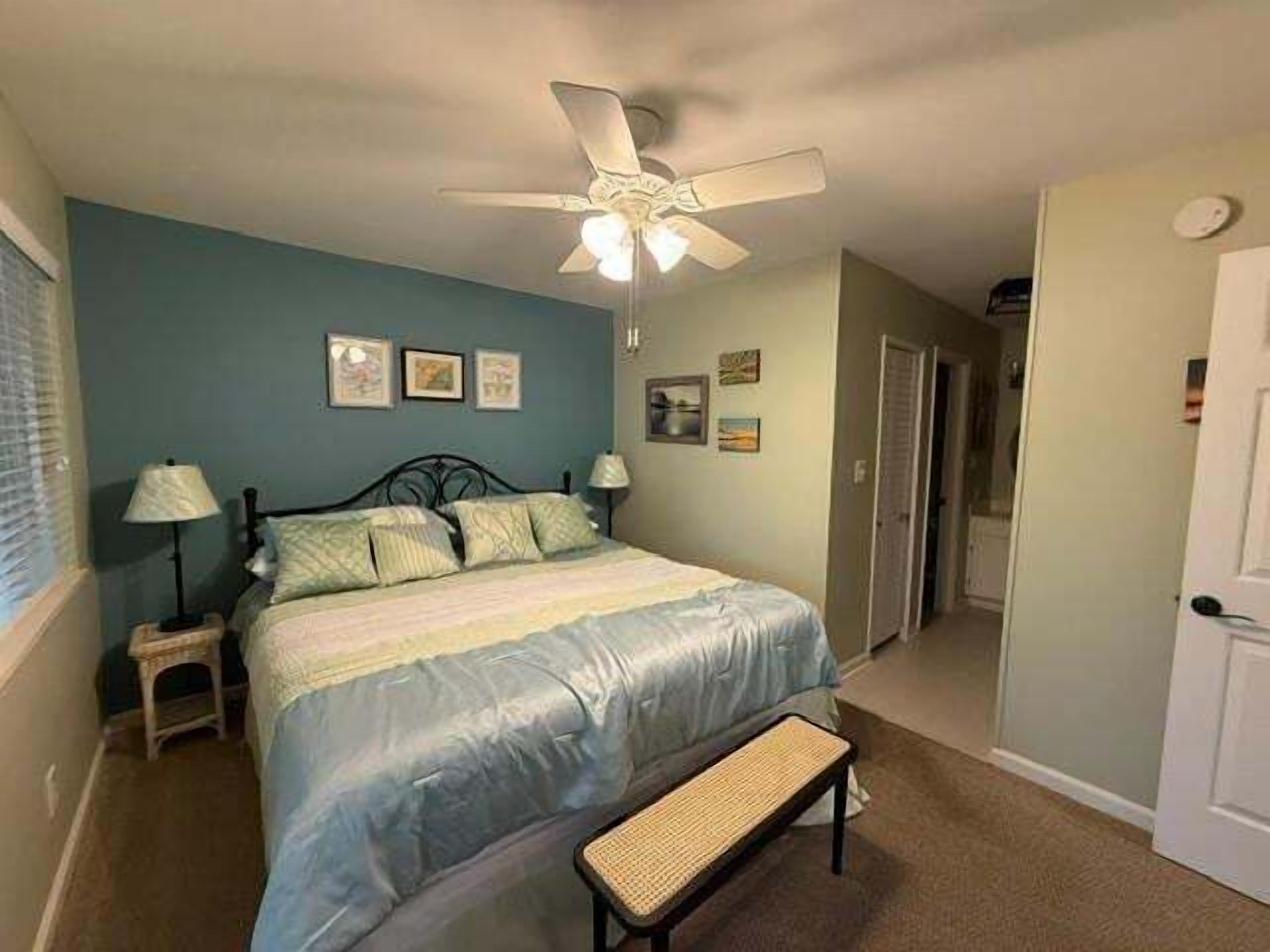 A bedroom with a bed, a ceiling fan, a bench, and a door.