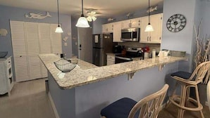 A kitchen with a granite countertop, white cabinets, a microwave, and a clock on the wall.
