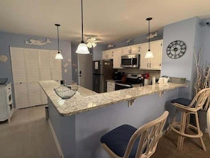 A kitchen with a granite countertop, white cabinets, a microwave, and a clock on the wall.