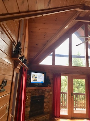 Interior de uma cabana de madeira com lareira de pedra, uma televisão montada acima e uma varanda com guarda-corpo de madeira.