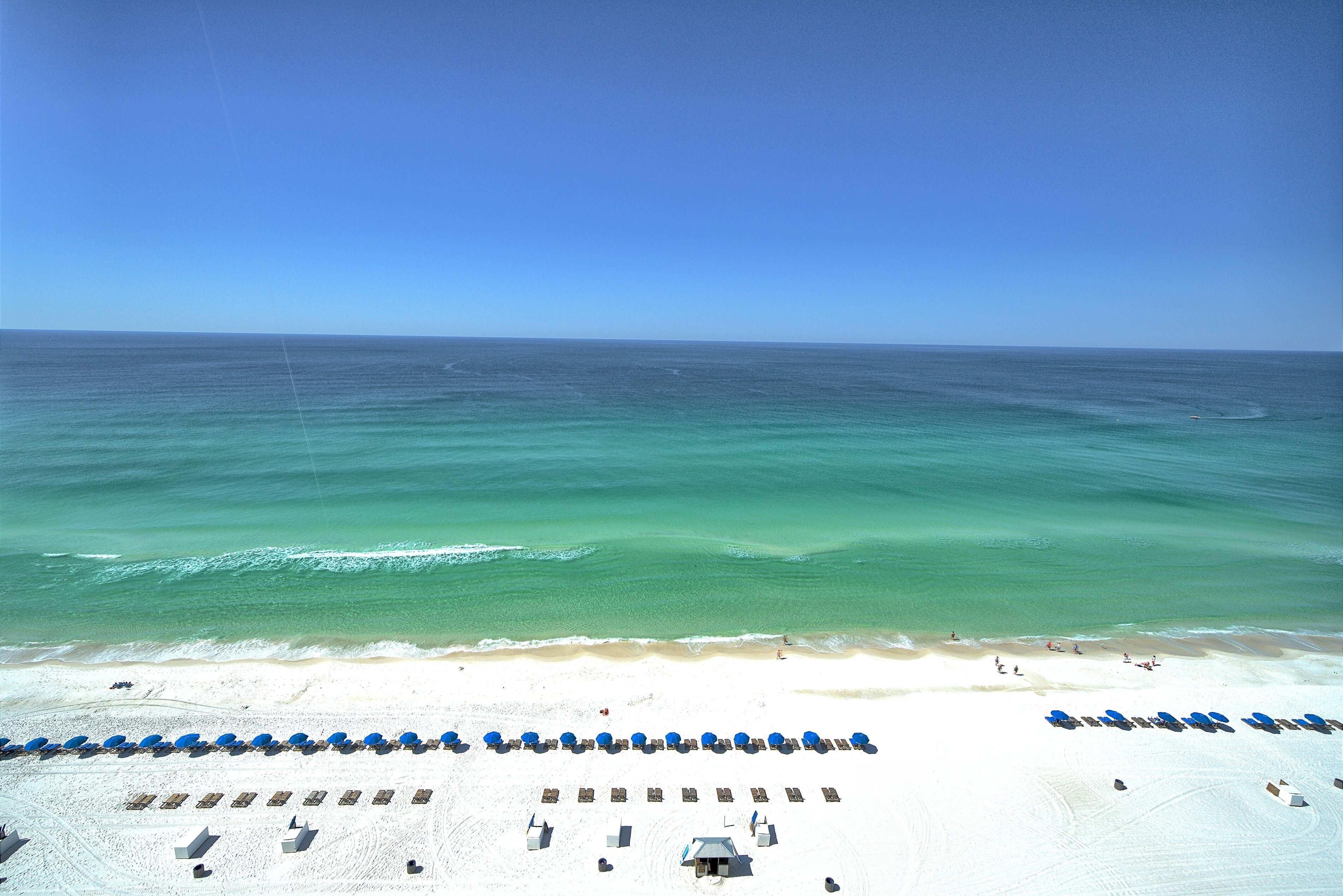 A beach with rows of blue umbrellas and sun loungers, a sandy beach, and the ocean.