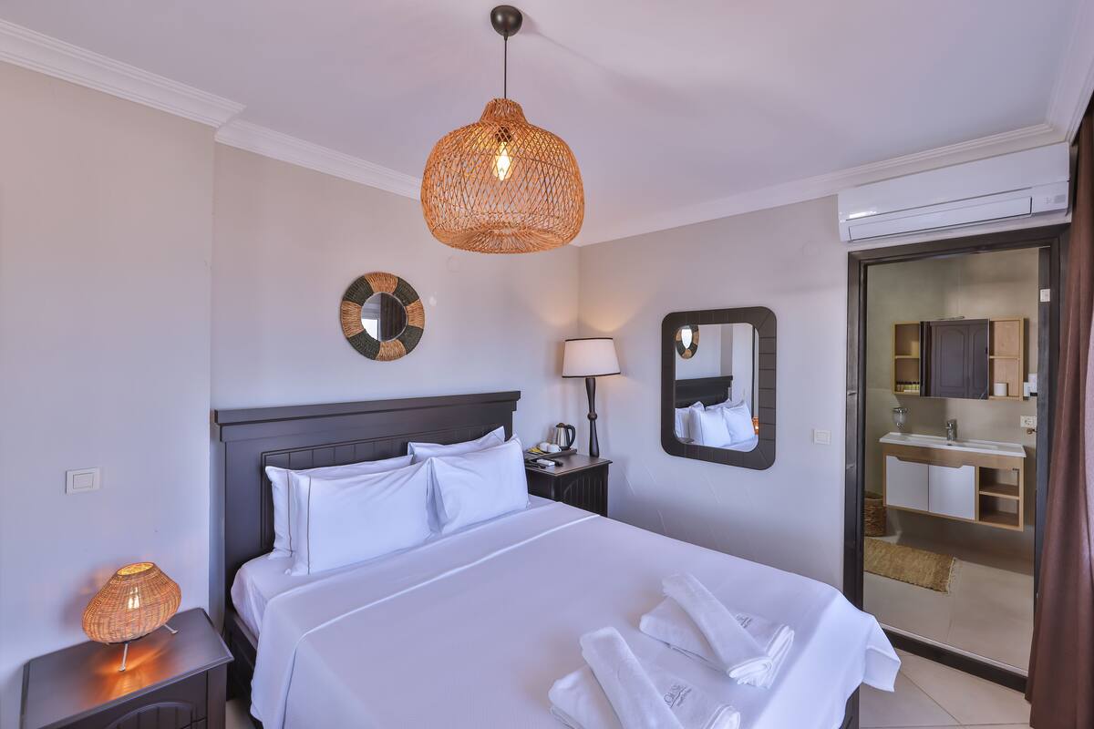 A hotel room with a bed, bedside lamp, wall clock, and a bathroom visible through a glass door.