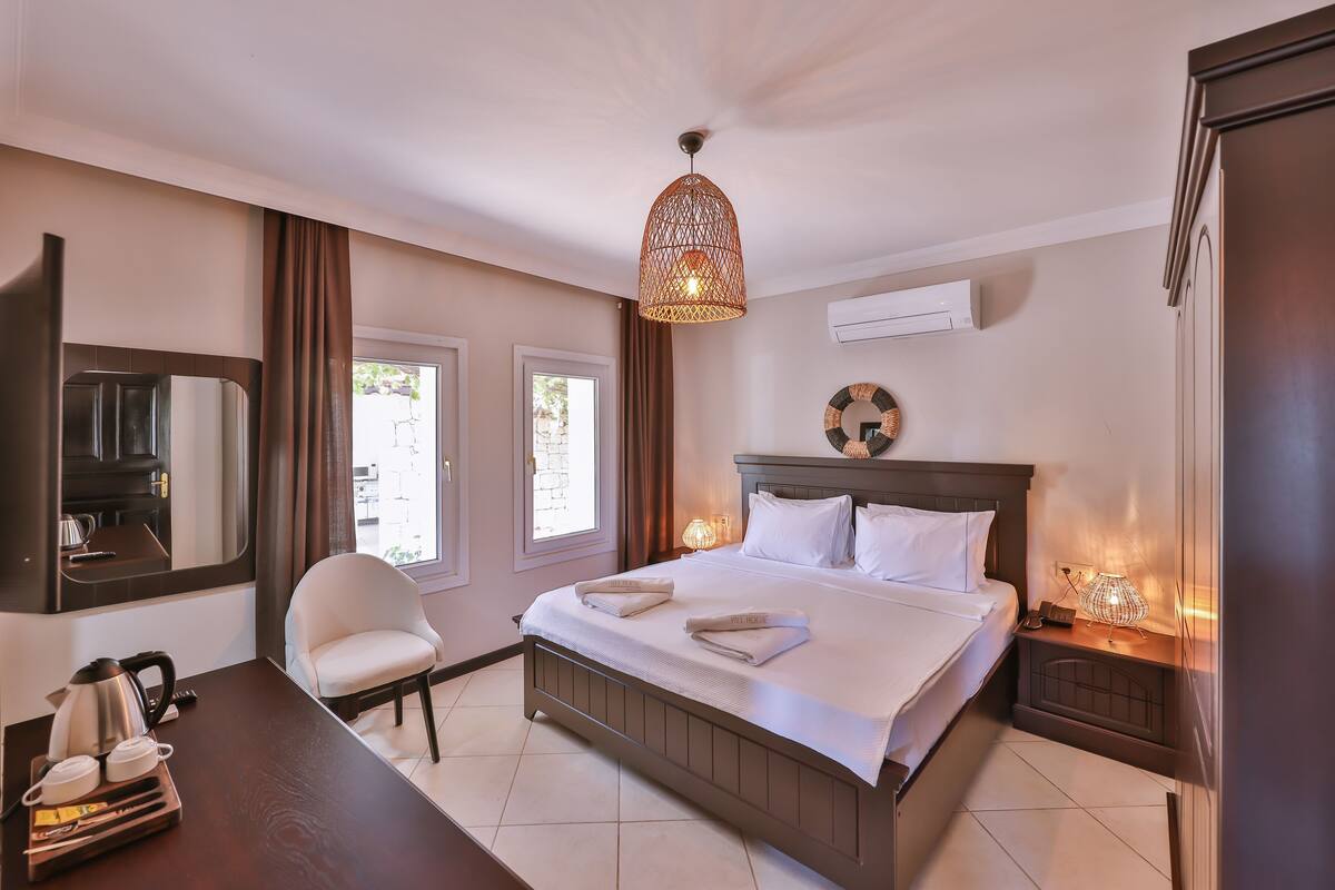 A hotel room with a bed, a chair, a desk with a kettle, a lamp, and a mirror.