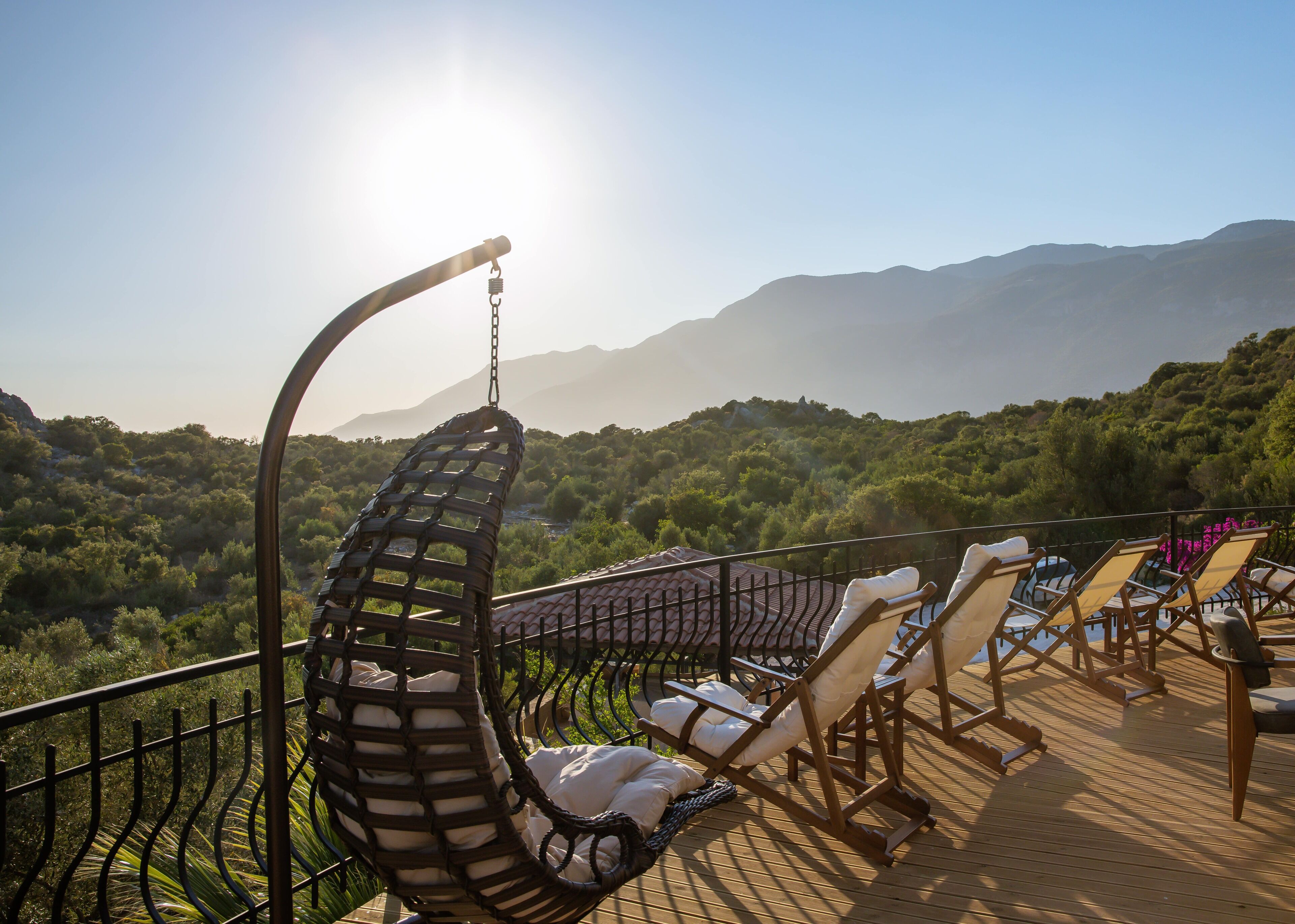 A sunlit outdoor seating area with lounge chairs and a hanging chair, overlooking a mountainous landscape.