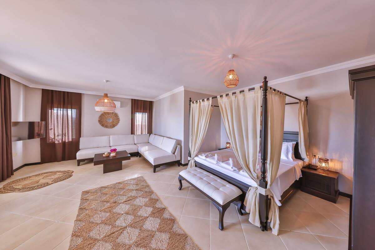 A spacious bedroom with a four-poster bed, a sitting area with a sofa and a coffee table, and a large window with curtains.