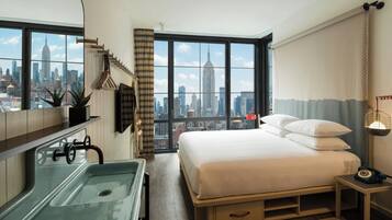 A hotel room with a large window, a bed, a bedside table, a nightstand, a sink, and a mirror.