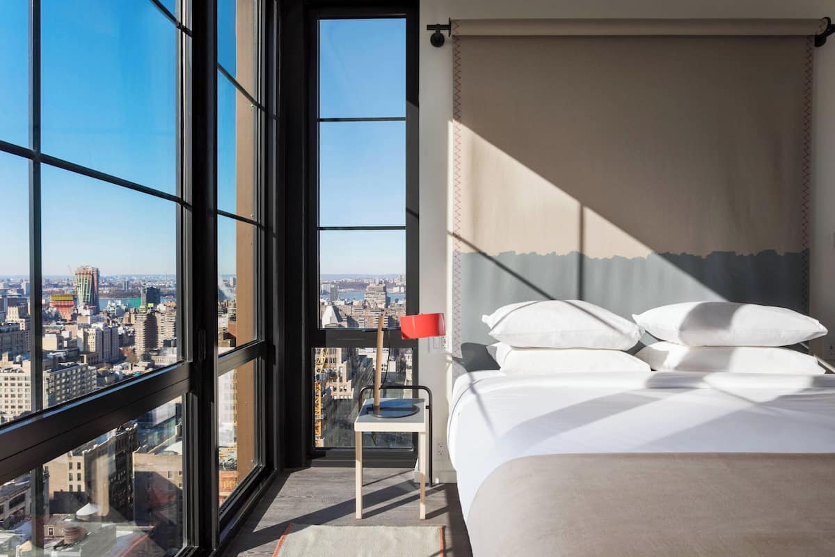 A hotel room with a large window offering a city view, a bed with white linens, and a red chair.