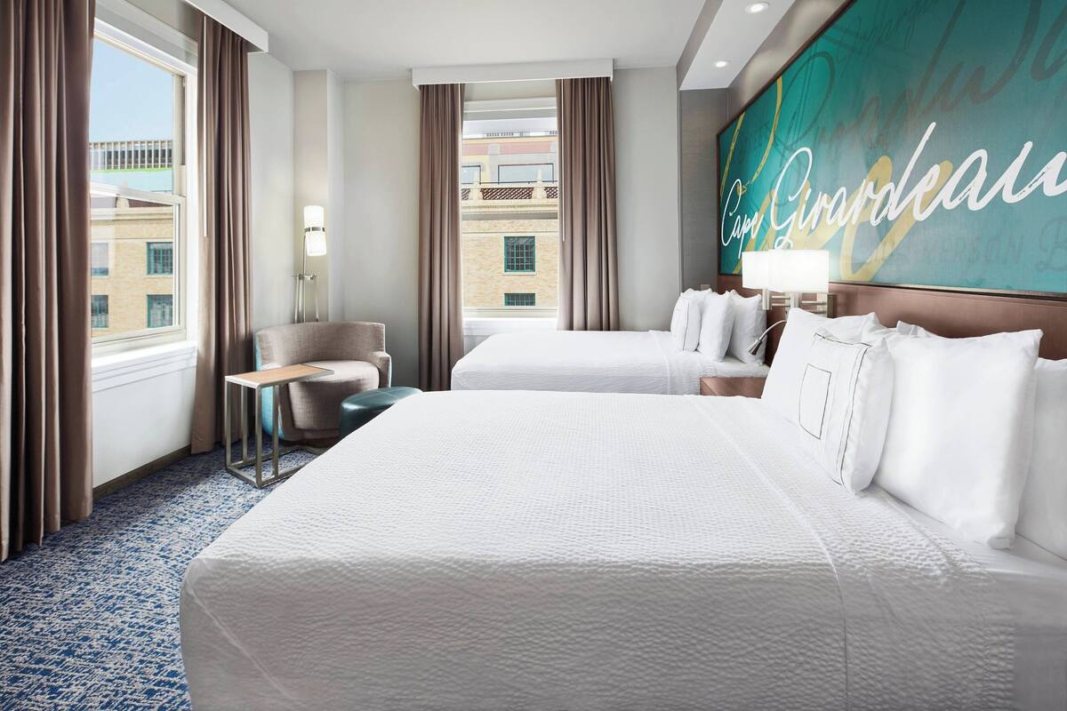 Ein Hotelzimmer mit zwei Betten, einem Schreibtisch, einem Stuhl und einem großen Fenster mit Vorhängen.