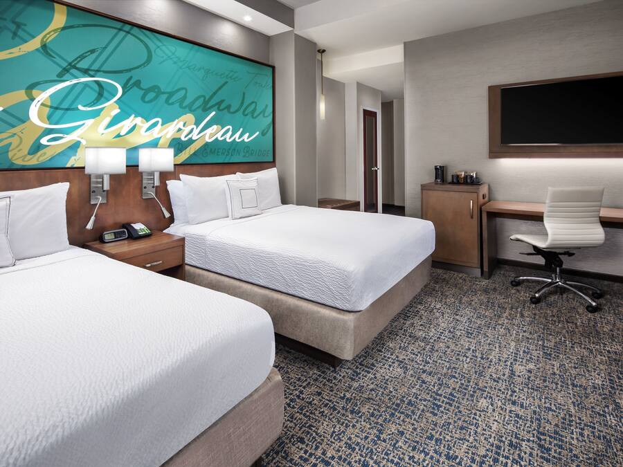 A hotel room with two beds, a desk, a chair, a TV, and a bathroom visible in the background.
