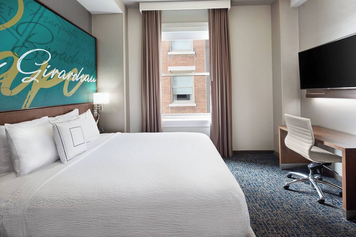 Ein Hotelzimmer mit einem großen Bett, einem Schreibtisch, einem Sessel, einem Fernseher und einem Fenster mit Vorhängen.
