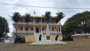 A building with a balcony, palm trees, and two cannons in front. - Pousada Água Viva (Fernando de Noronha)