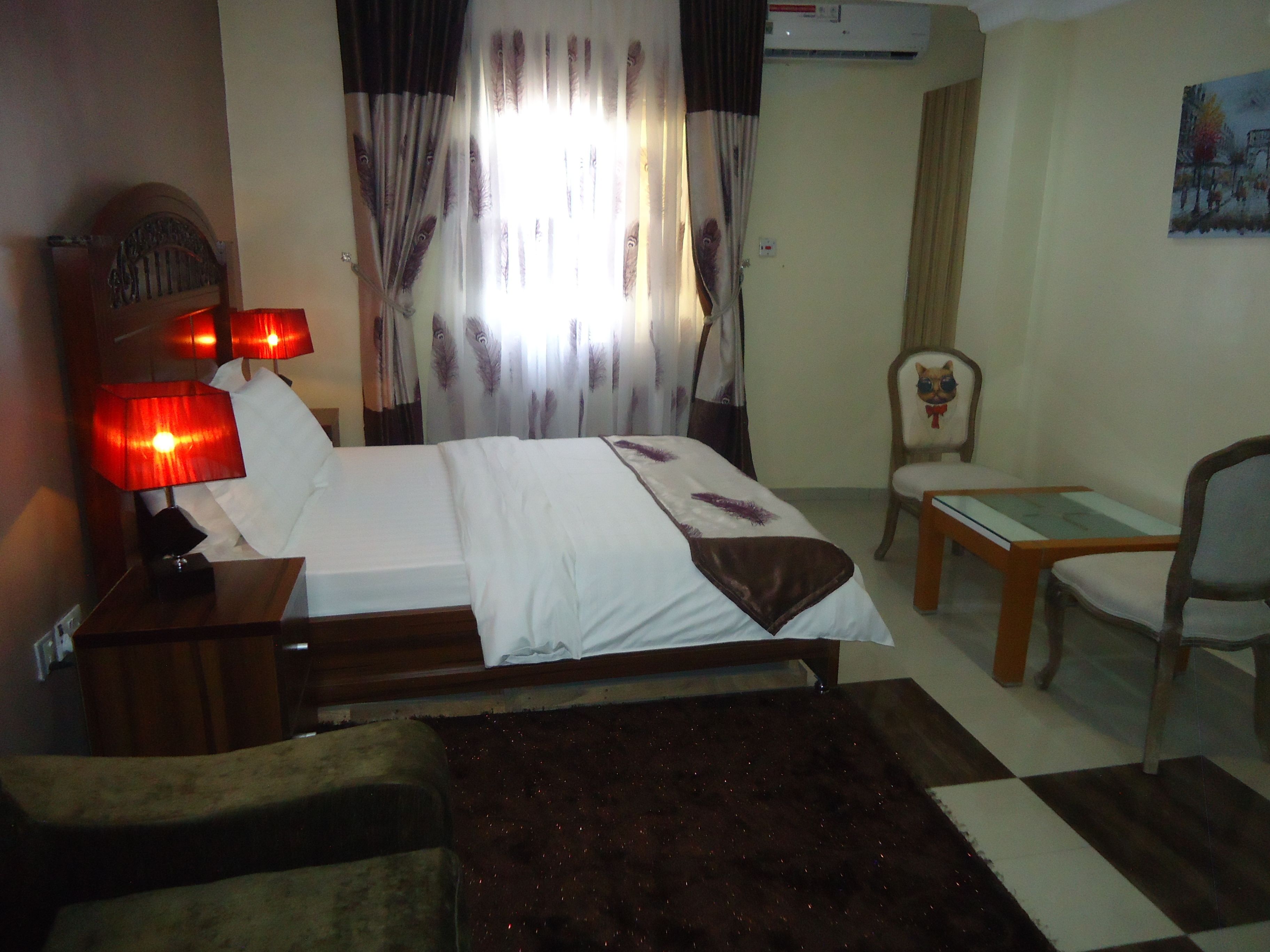 A hotel room with a bed, a chair, a small table, a nightstand with a lamp, and a painting on the wall.