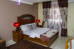Une chambre à coucher avec un lit en bois, deux lampes de chevet, une table de nuit, une grande fenêtre avec des rideaux et un sol recouvert d’une moquette.