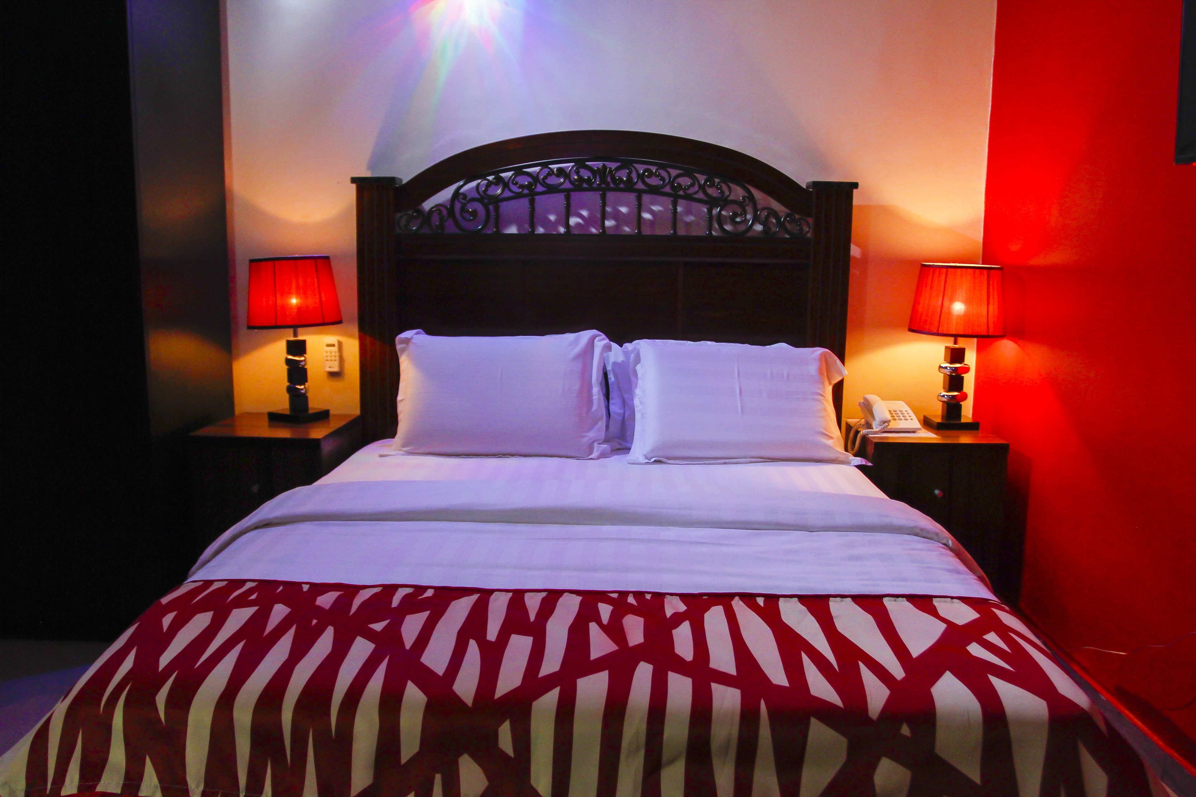 A neatly made bed with white linens and a patterned bedspread, two bedside tables with lamps, and a red accent wall.