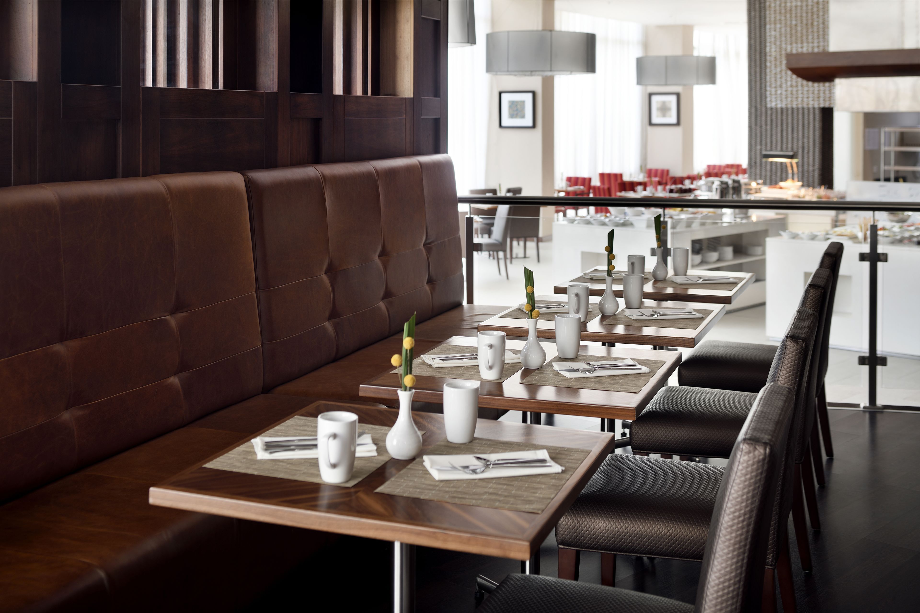 A restaurant with a long wooden table set with white plates, silverware, and vases with flowers. There is a brown leather booth and dark chairs.