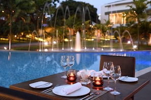A dining table set for two with glasses and candles, overlooking a pool with water fountains and a building in the background. - Accra Marriott Hotel (Accra)