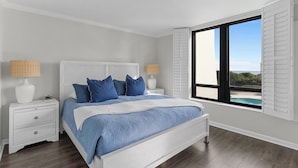 A bedroom with a bed, nightstands, and a window with shutters. - Stunning pool and ocean views await you in this beautiful, convenient condo. (Wrightsville Beach)