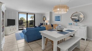 A modern living room with a blue sofa, white dining table, and a large window overlooking a balcony. - Stunning pool and ocean views await you in this beautiful, convenient condo. (Wrightsville Beach)