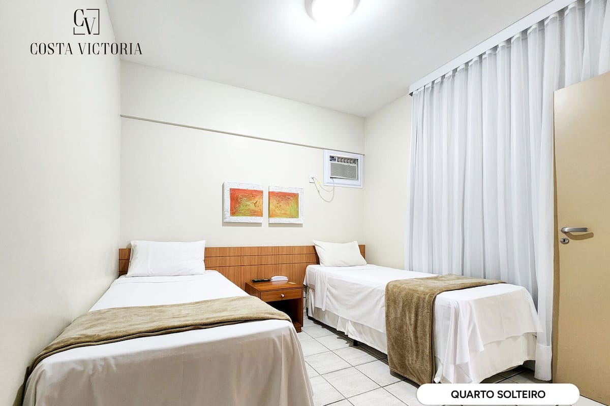 A hotel room with two single beds, a wooden headboard, a small bedside table, a nightstand, and a window with white curtains.