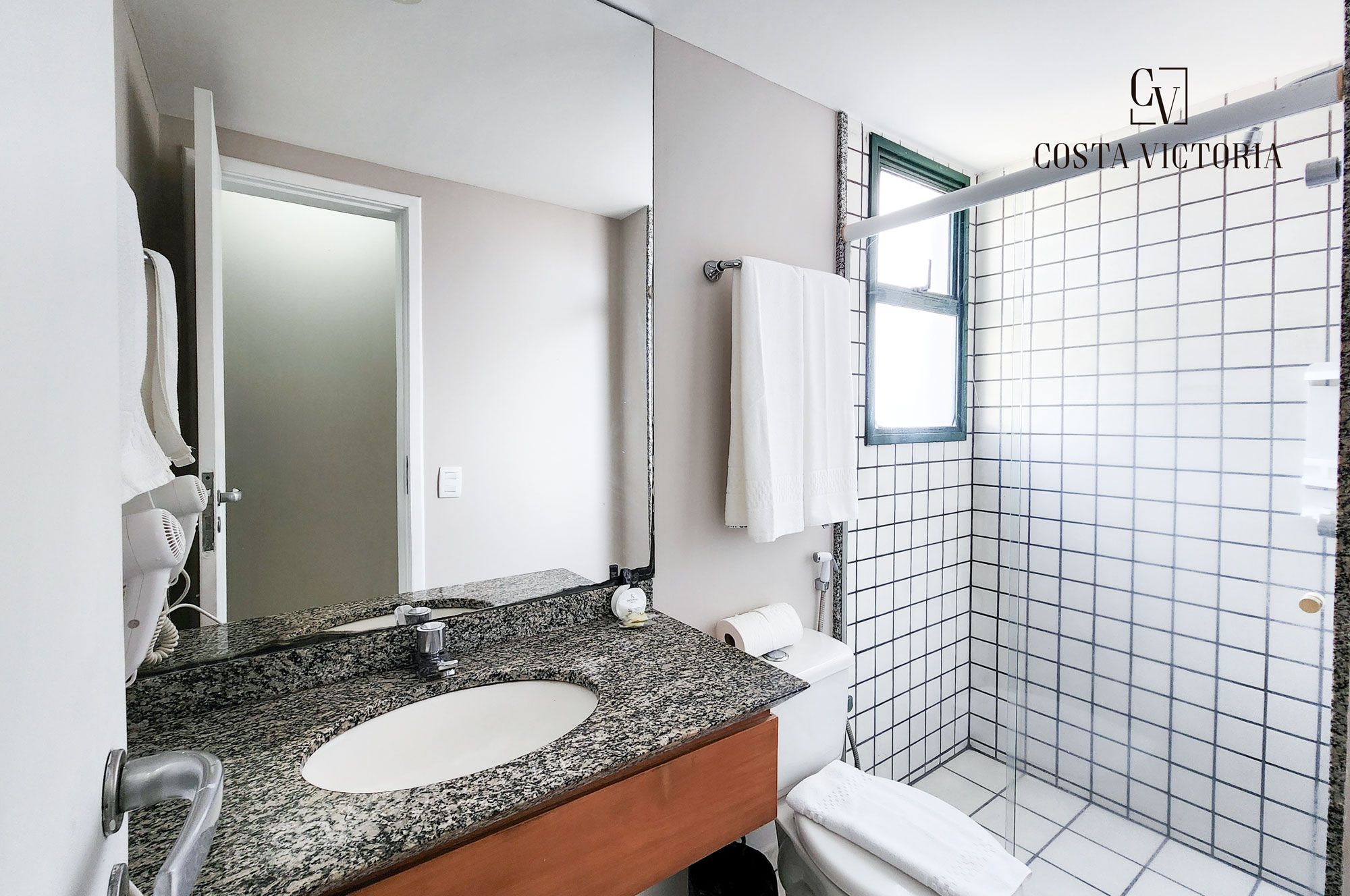 A bathroom with a granite countertop, a white sink, a mirror, a towel hanging on the wall, and a shower area with tiled walls.