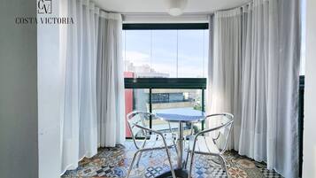 Un balcón con suelo estampado, una mesa redonda y dos sillas. El balcón tiene cortinas blancas y vistas a edificios.
