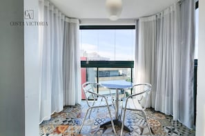 Un balcón con suelo estampado, una mesa redonda y dos sillas. El balcón tiene cortinas blancas y vistas a edificios.