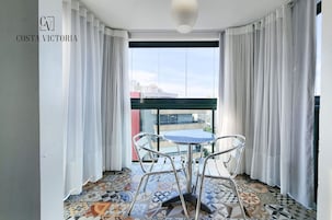 Un balcón con suelo estampado, una mesa redonda y dos sillas. El balcón tiene cortinas blancas y vistas a edificios.