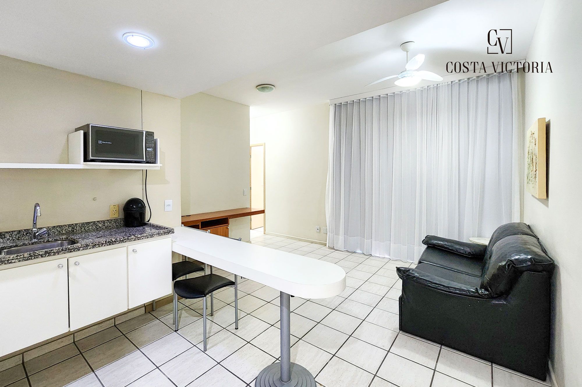 Uma cozinha compacta com micro-ondas, pia e bancada. Há uma mesa branca com uma cadeira preta no centro do quarto. Um sofá de couro preto está encostado na parede, e um ventilador de teto é visível.