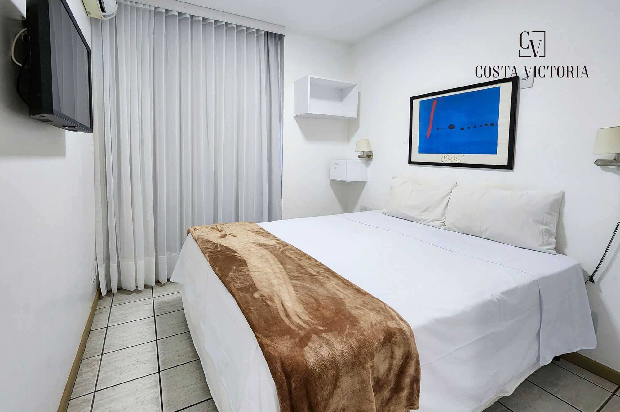 Quarto de hotel com cama, televisão, janela com cortinas, prateleira na parede e um quadro emoldurado.