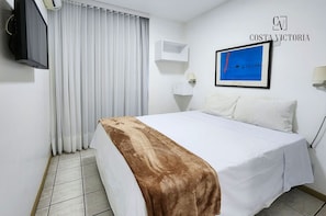 A hotel room with a bed, a TV, a window with curtains, a wall-mounted shelf, and a framed picture. - Costa Victoria Residence (Vitoria)