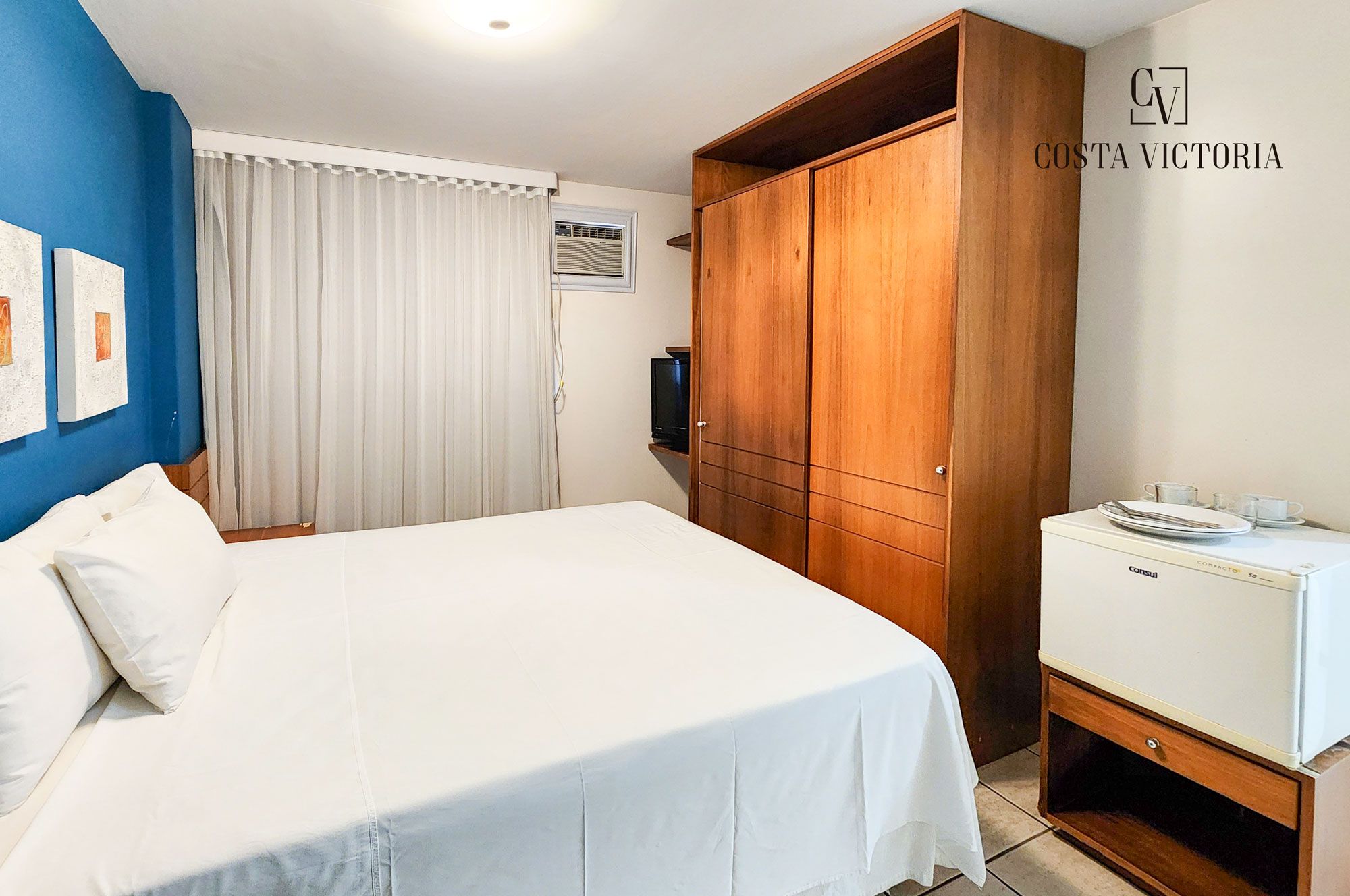 A hotel room with a large bed, wooden wardrobe, and a compact kitchenette.